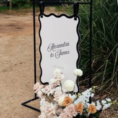 a sign that is sitting on the ground with flowers in front of it and some bushes behind it