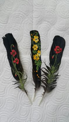 three feathers with flowers painted on them sitting on a white quilted bed spreader