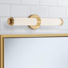 a bathroom mirror sitting next to a wall mounted light on a white tiled wall with gold trim