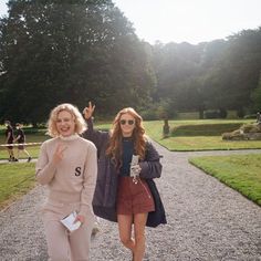 two young women walking down a path in the middle of a park, one holding her hand up