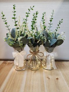 three glass vases with flowers and greenery in them on a wooden table top