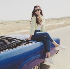 a woman sitting on the hood of a blue car next to a desert road and wearing sunglasses