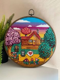 a cross stitch picture hanging on a wall next to a potted plant with a house in the background