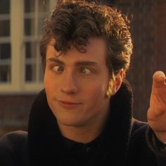 a young man with curly hair and green eyes pointing to the side while wearing a black coat