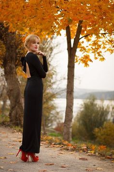 a woman in black dress and red shoes standing by trees with yellow leaves on the ground