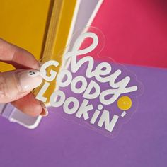 a hand holding a sticker that says hey goody looking in white on it