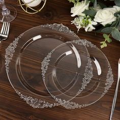 two clear plates on a wooden table with silverware and white flowers in the background