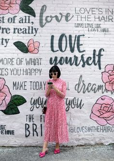 a woman standing in front of a white brick wall with pink flowers and writing on it