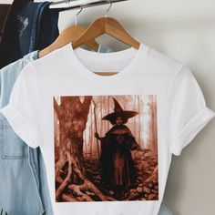 This adult unisex short sleeve tee features a dark and spooky tinted image of an afro witch in the woods. Crafted with soft cotton and a quality print, this tee will become your well-loved fall favorite. It's a great top to celebrate the spirit of Halloween, but the illustration works year round as well. Its ribbed knit collars, taping on the shoulders, and dual side seams ensure a better fit over time and maintain the garment's shape. Made with 100% cotton, this tee offers a comfortable retail fit and light fabric weighing 4.2 oz/yd² (142 g/m²). With its tear away label and true-to-size fit, our witchy tee is perfect for those who want style, comfort, and a magical design. Available in sizes XS-5XL. Easily customizable upon request. Shop The Trini Gee for more: https://www.etsy.com/shop/t Unisex Fall Short Sleeve T-shirt, Unisex Graphic Print T-shirt For Fall, Fitted Band Merch T-shirt For Fall, Unisex Short Sleeve T-shirt For Fall, Fall Short Sleeve T-shirt, Unisex Halloween Graphic Print Top, Unisex Short Sleeve Halloween T-shirt, Unisex Graphic Print Top For Halloween, Unisex Cotton Halloween T-shirt