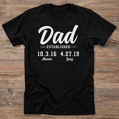 a black t - shirt with the words dad established and date printed on it, sitting on a wooden surface