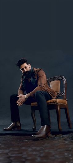 a man sitting on top of a wooden chair in front of a black wall with his hand on his knees