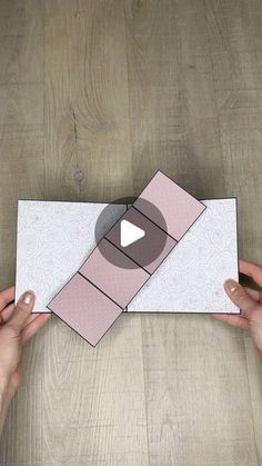 two hands holding an origami piece on top of a wooden table with a video playing button