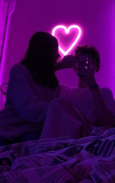 a man and woman sitting on a bed in front of a neon heart