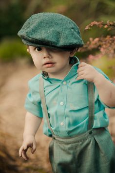 a young boy wearing a green hat and suspenders with his hands on his hips