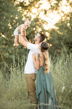 a man holding a baby up to his face while standing in tall grass with trees behind him