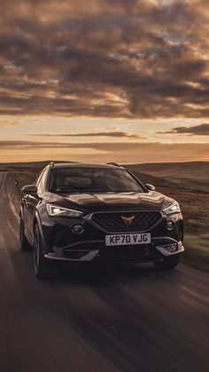 the front end of a black car driving down a country road at sunset with clouds in the background