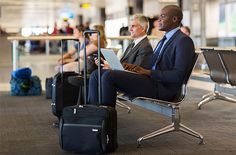 two men in suits sitting on chairs with their luggage and looking at the screen while waiting