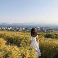 a woman in a white dress walking through tall grass with the city in the background