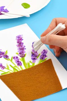 someone is painting flowers on paper with white paint and purple felt, while another hand holds a crochet hook