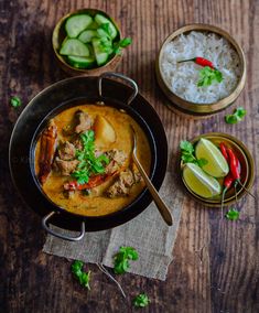 two bowls filled with food on top of a wooden table next to rice and cucumbers