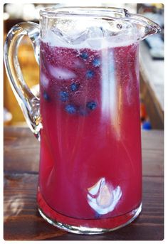 a pitcher filled with blueberries and ice on top of a wooden table next to a window