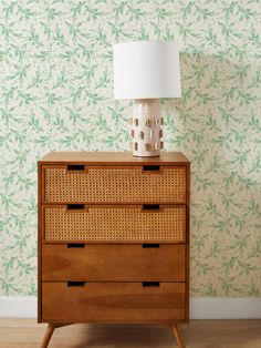 a wicker dresser with a lamp on top and green wallpaper behind the drawers