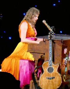 a woman in an orange dress sitting at a table with a guitar and singing into a microphone