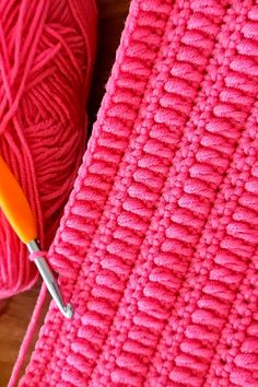 a pink crochet blanket next to a ball of yarn with an orange knitting needle