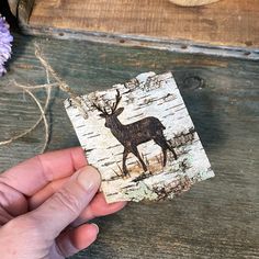 a hand holding a piece of wood with a deer on it and flowers in the background