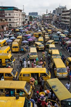 a large group of yellow buses parked in a parking lot next to tall buildings and people with umbrellas