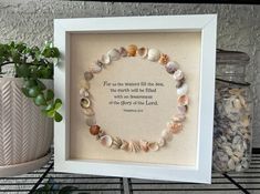 a white framed frame with shells and a quote on it next to a potted plant