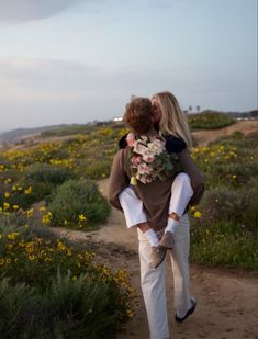 a man carrying a woman on his back down a dirt path near flowers and grass