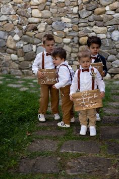 three young boys dressed in brown and white holding wooden signs with writing on them, standing next to a stone wall