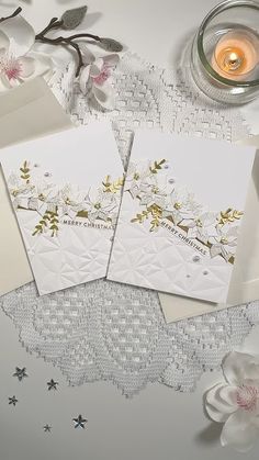 three white cards with gold foil on them next to some flowers and a lit candle