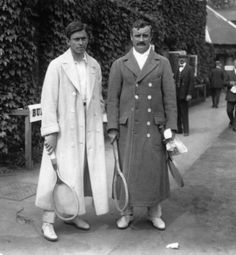 two men in long coats holding tennis racquets while standing next to each other