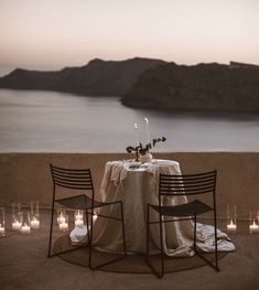 two chairs and a table set up with candles on the ground in front of water