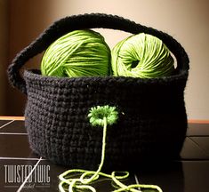 two balls of yarn are in a black crochet basket with green thread on the floor