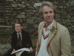 two men sitting next to each other in front of a stone wall and holding papers