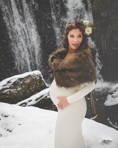 a pregnant woman standing in front of a waterfall wearing a fur stole with flowers on her head