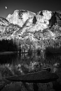 black and white photograph of the moon setting over a mountain range with a lake in front of it