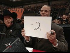 a man holding up a sign in front of an audience at a wrestling match with another man wearing a cowboy hat