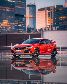 a red car parked in front of some tall buildings with its reflection on the ground