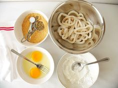 three bowls with noodles, eggs and flour in them on a white tablecloth next to two silver spoons