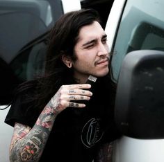 a man with long hair and piercings leaning against a white car door looking at the mirror