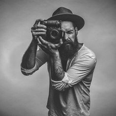 a man with a beard holding a camera up to his face while wearing a hat