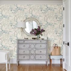 a dresser with flowers on it in front of a wallpapered mirror and chair