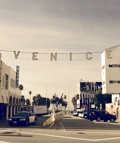 a man walking down the street carrying a surfboard under an arch that reads venice