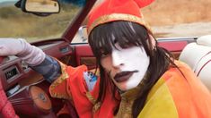 a woman with makeup on sitting in the driver's seat of a car wearing an orange and yellow costume