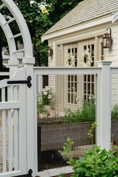 a white fence with an arched top and gate in front of a house, surrounded by greenery