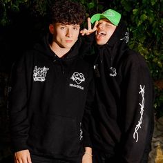 two young men standing next to each other wearing black hoodies and green caps on their heads